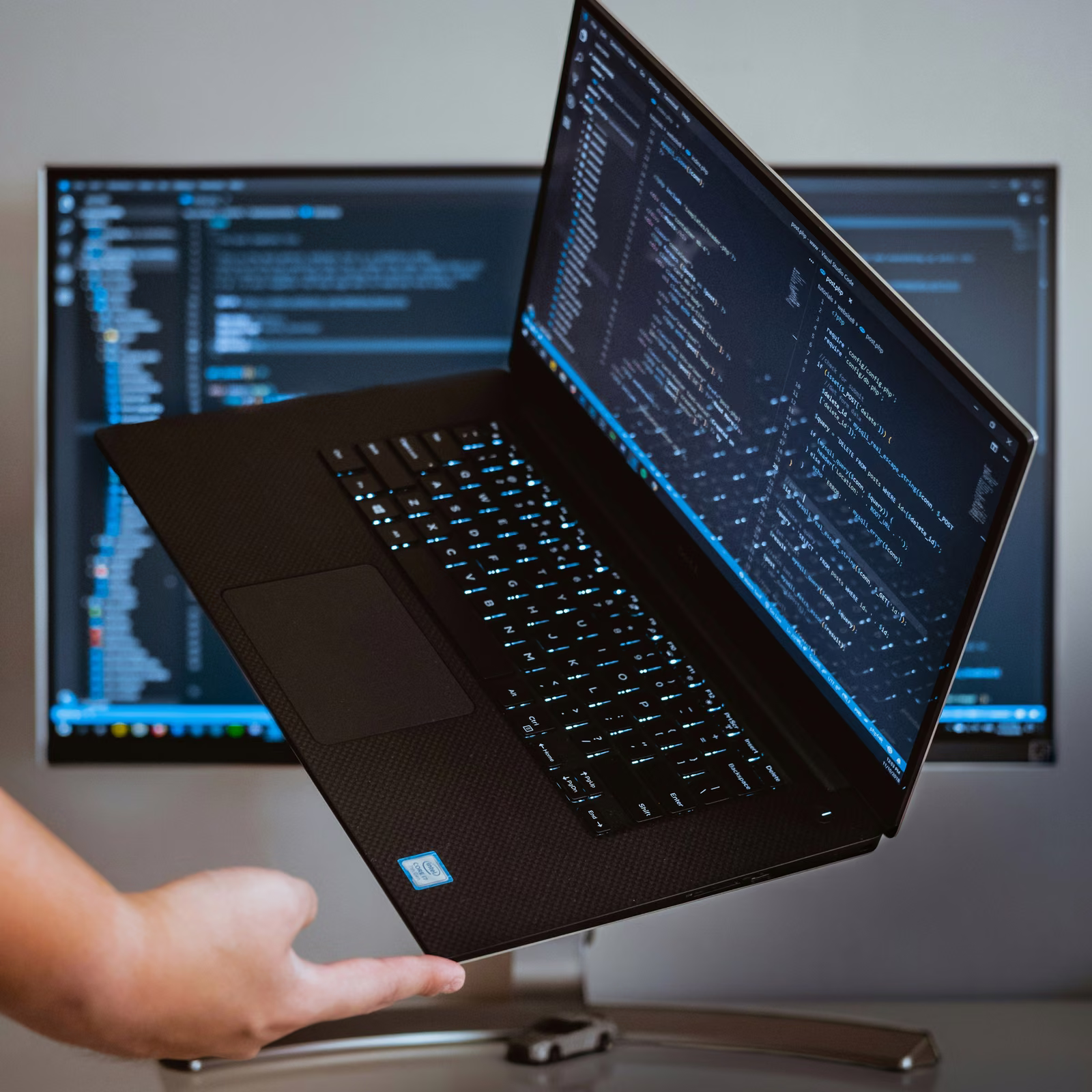 Developer holding up a laptop showing code in front of a monitor with more code on screen