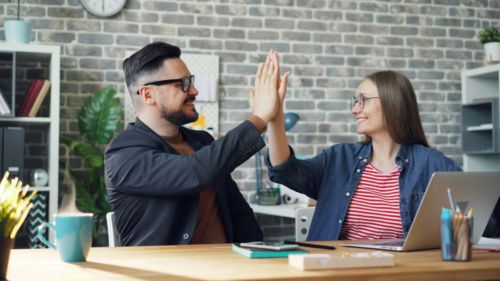 Two smiling coworkers sharing a high five at a desk with a laptop, celebrating a resolved issue